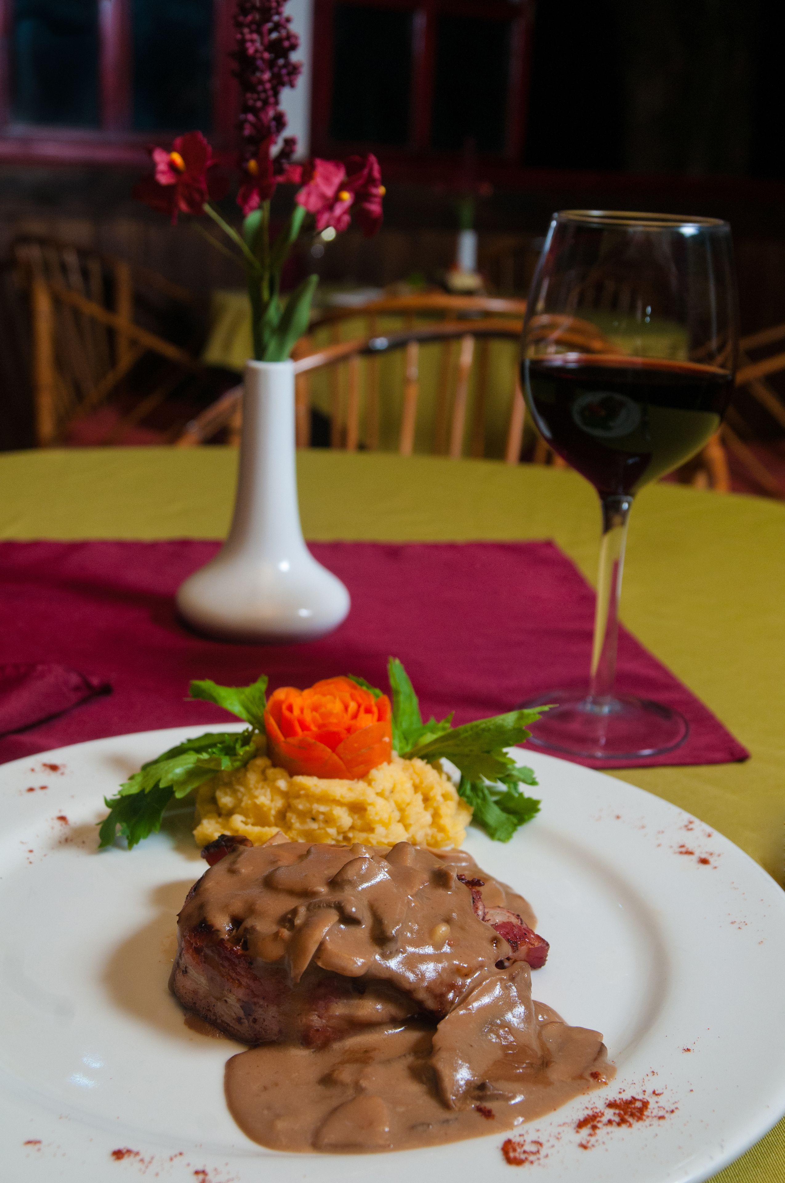 Una mesa de comedor con un plato de comida, una copa de vino tinto y un florero con flores.
