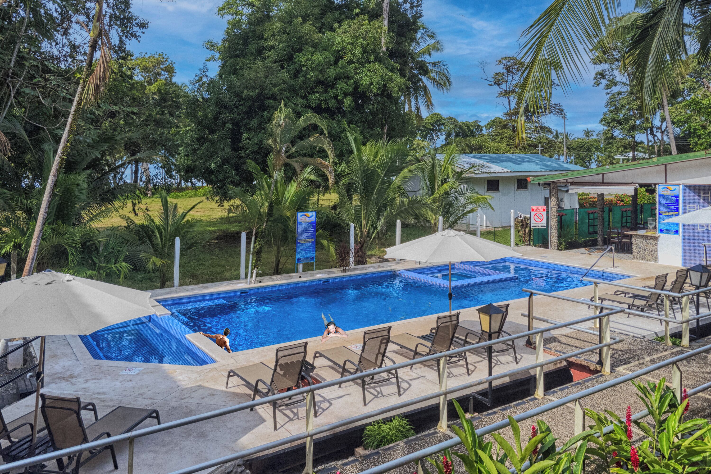 A swimming pool with lounge chairs and umbrellas, surrounded by greenery and a building with a green roof.