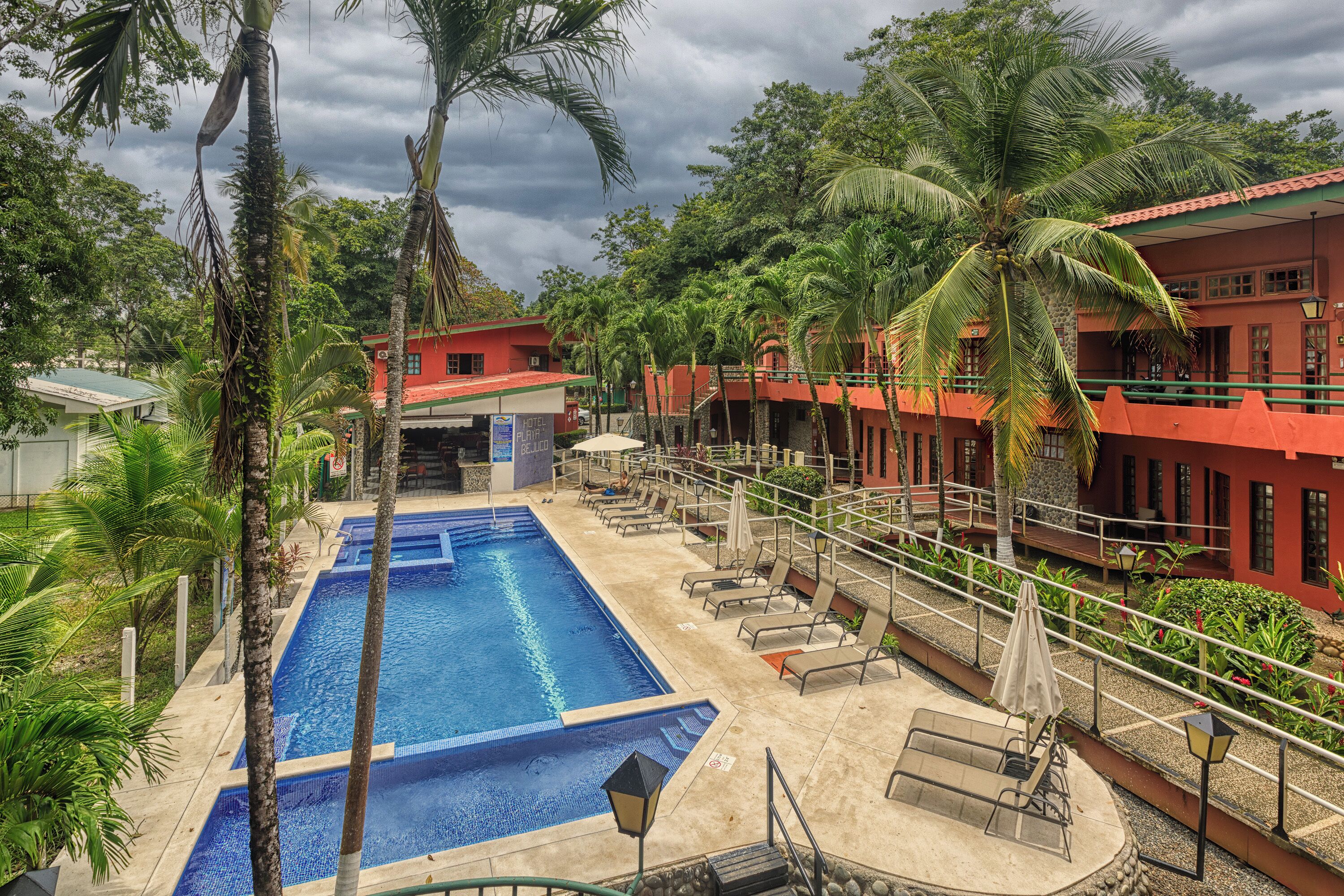 A swimming pool with lounge chairs and umbrellas, surrounded by palm trees and a red building.
