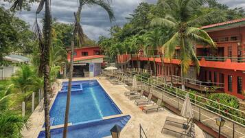 A swimming pool with lounge chairs and umbrellas, surrounded by palm trees and a red building.