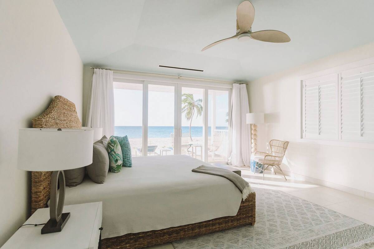 A bedroom with a bed, a ceiling fan, a window with curtains, a beach view, and a chair by the window.