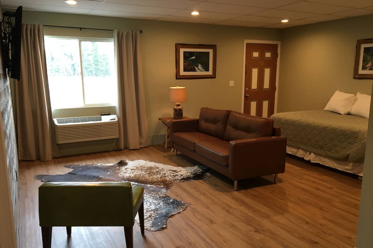 A room with a wooden floor, a brown leather sofa, a small table with a lamp, a bed with white bedding, and a large window with curtains.