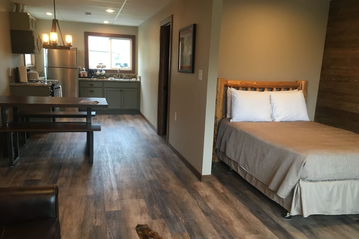 A modern bedroom with a wooden bed, a dining area with a bench, and a kitchen in the background.