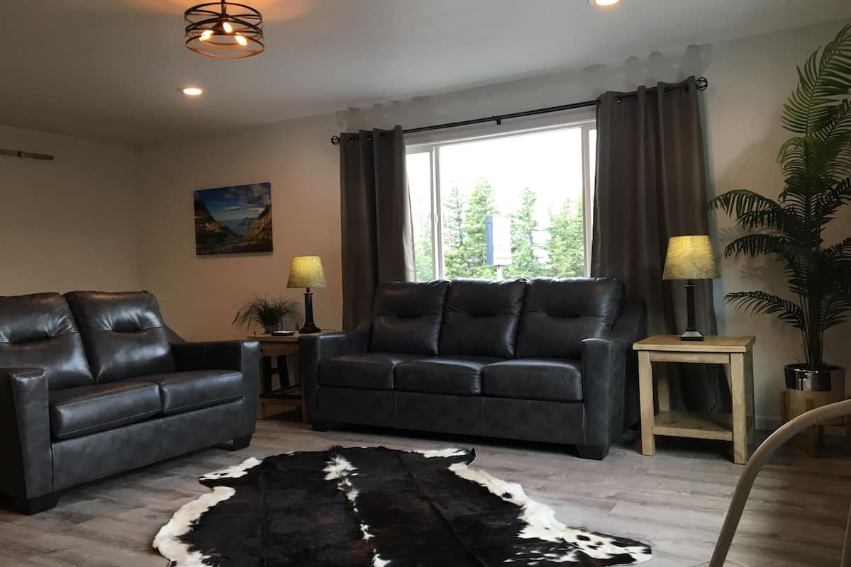 A living room with two leather sofas, a black and white rug, a wooden coffee table, a potted plant, and a window with curtains.