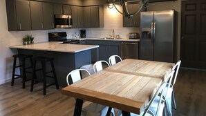 A modern kitchen with dark cabinets, a central island, a stainless steel refrigerator, and a wooden dining table with white chairs. - Glacier Basecamp Lodge (Columbia Falls)