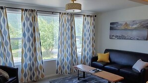 A living room with a sofa, a coffee table, and a rug. There are curtains on the windows, and a painting on the wall. - Glacier Basecamp Lodge (Columbia Falls)