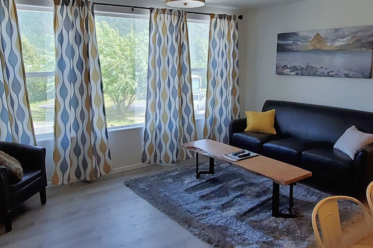 A living room with a sofa, a coffee table, and a rug. There are curtains on the windows, and a painting on the wall.