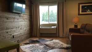 A room with a wooden wall, a mounted TV, a leather couch, a green chair, a fur rug, and a window with curtains. - Glacier Basecamp Lodge (Columbia Falls)