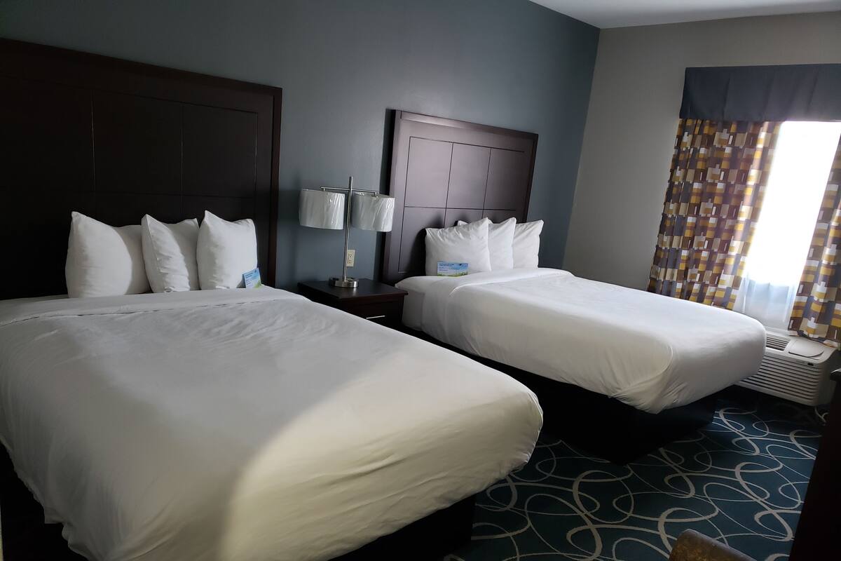 A hotel room with two beds, a nightstand with a lamp, a headboard, and a window with curtains.