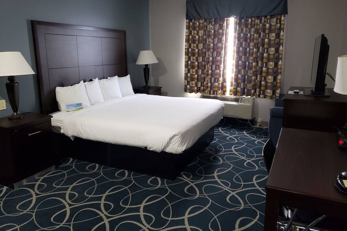 A hotel room with a large bed, two bedside tables with lamps, a desk with a chair, a window with patterned curtains, and a wall-mounted air conditioning unit.