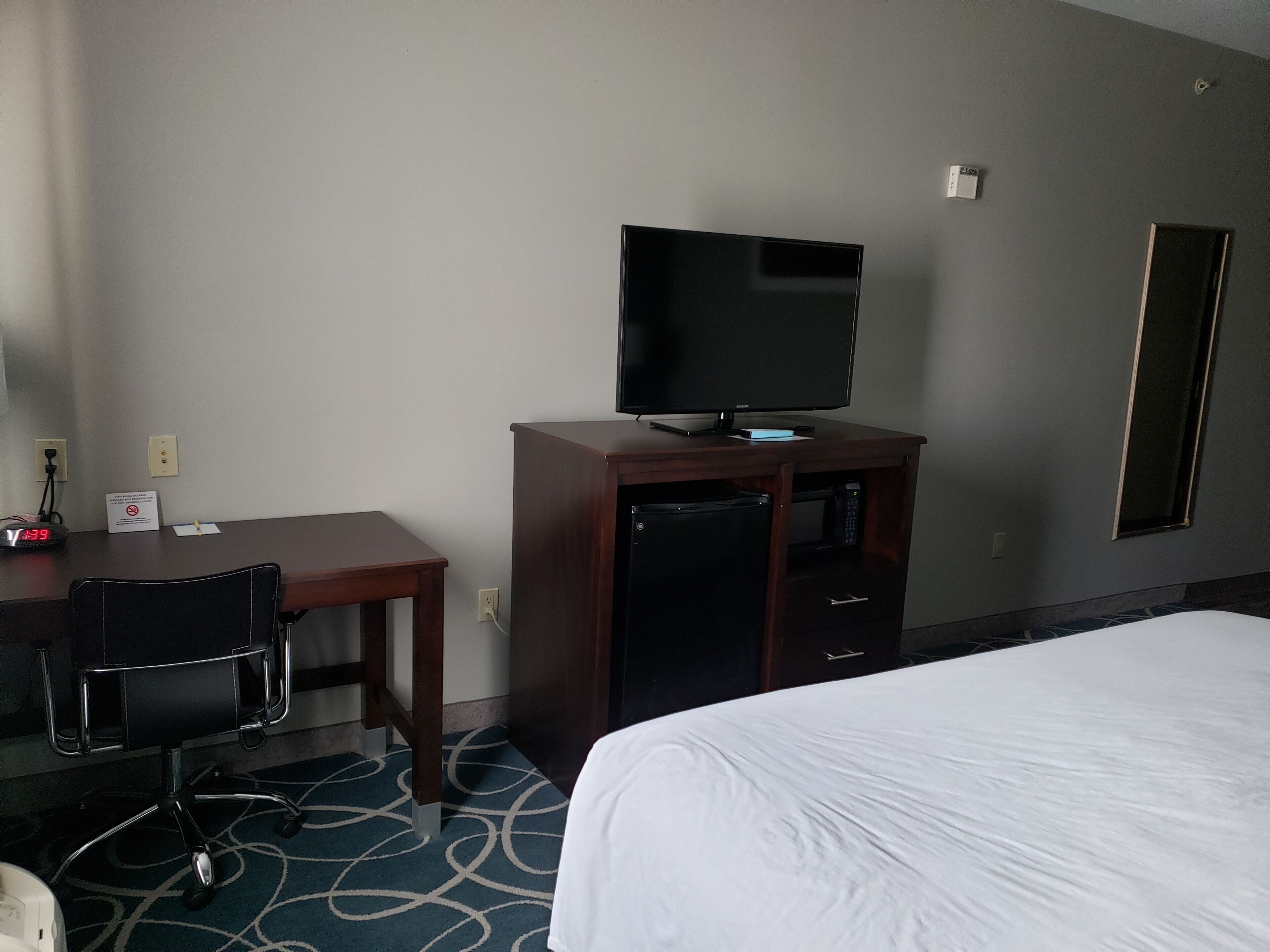 A hotel room with a bed, a desk with a chair, a television on a cabinet, and a mirror on the wall.