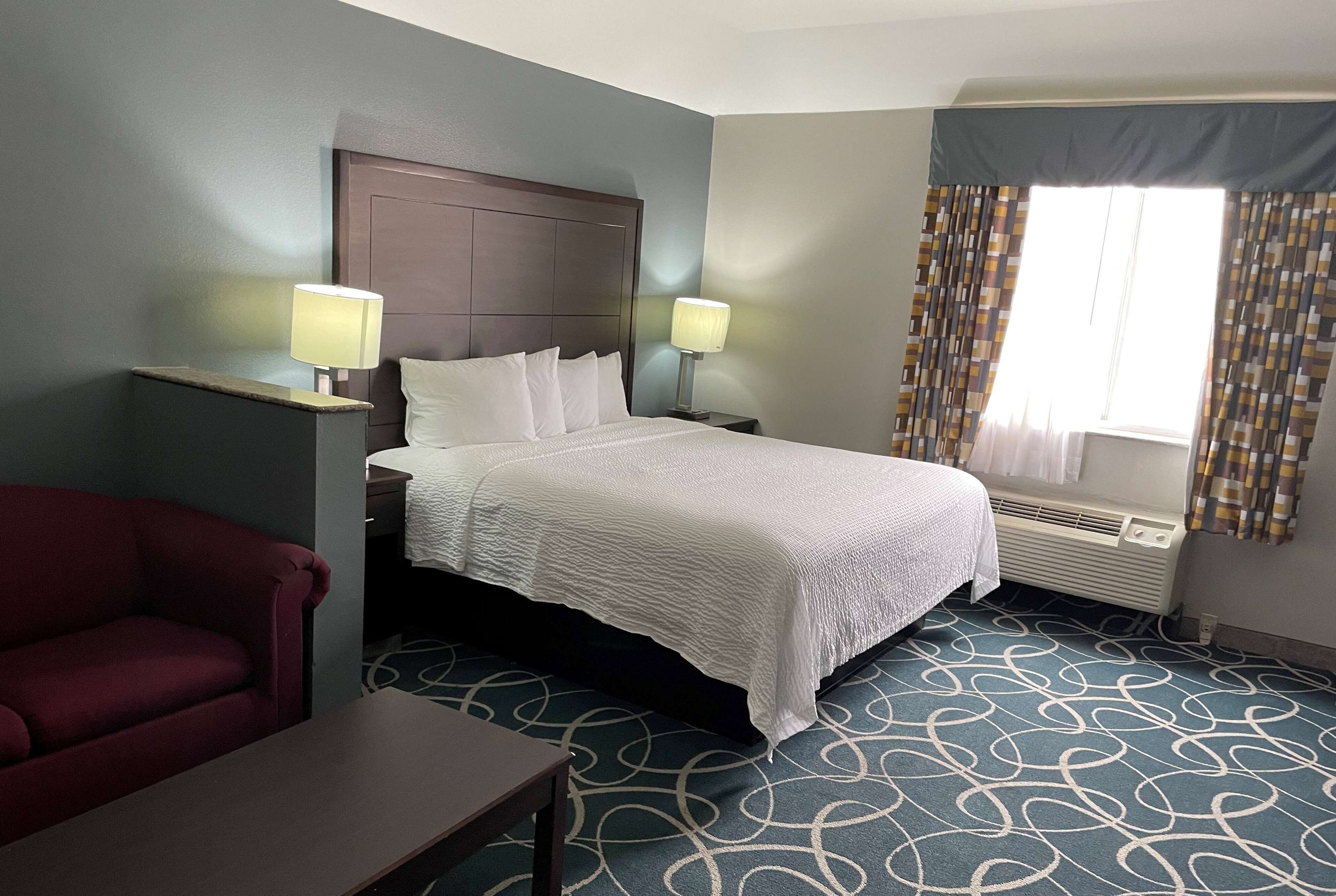 A hotel room with a bed, two bedside lamps, a red armchair, a small table, patterned carpet, and a window with curtains.