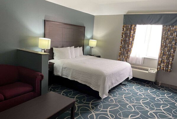 Quarto de hotel com cama, duas luminárias de cabeceira, poltrona vermelha, mesinha, carpete estampado e janela com cortinas.