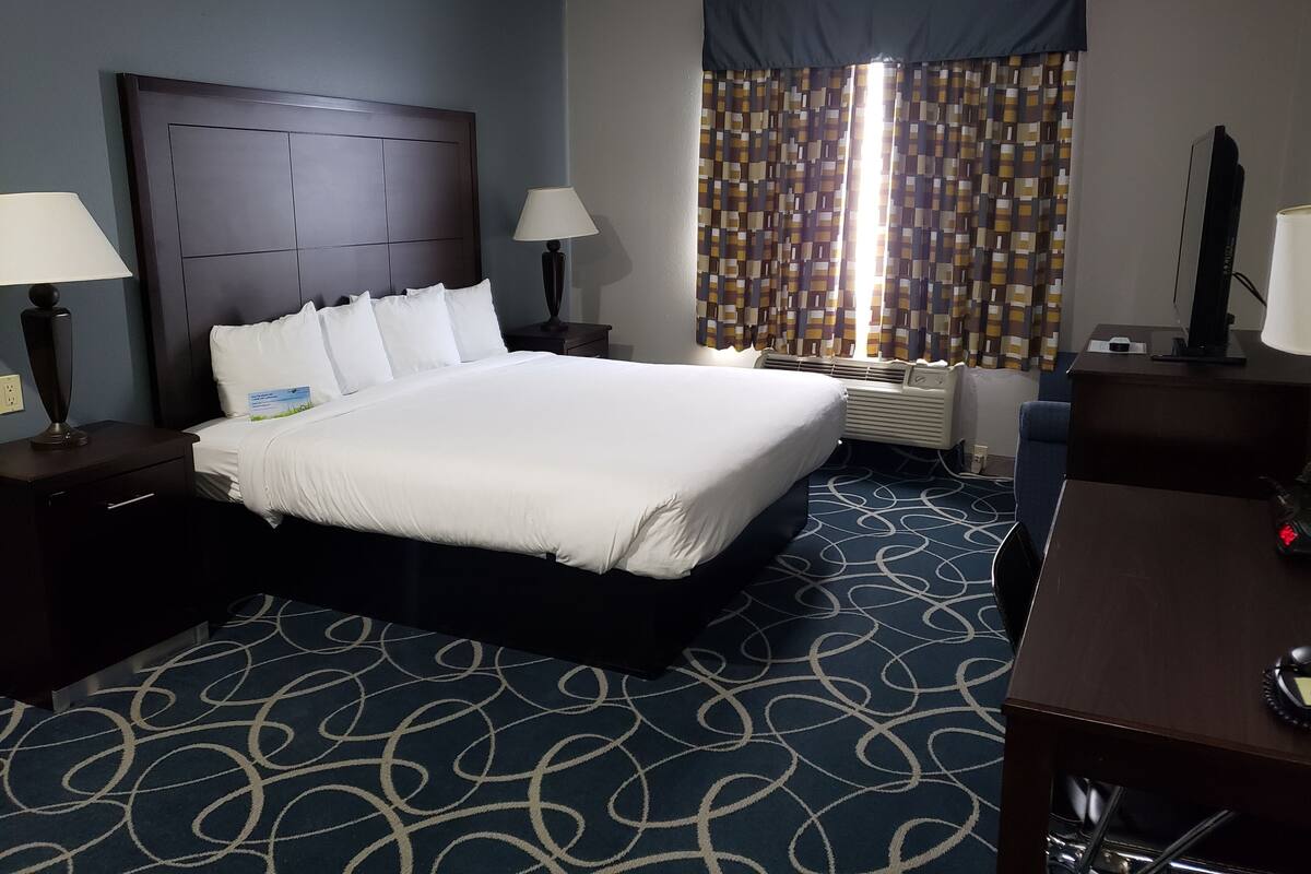 A hotel room with a large bed, two bedside tables with lamps, a desk with a chair, a window with patterned curtains, and a wall-mounted air conditioning unit.