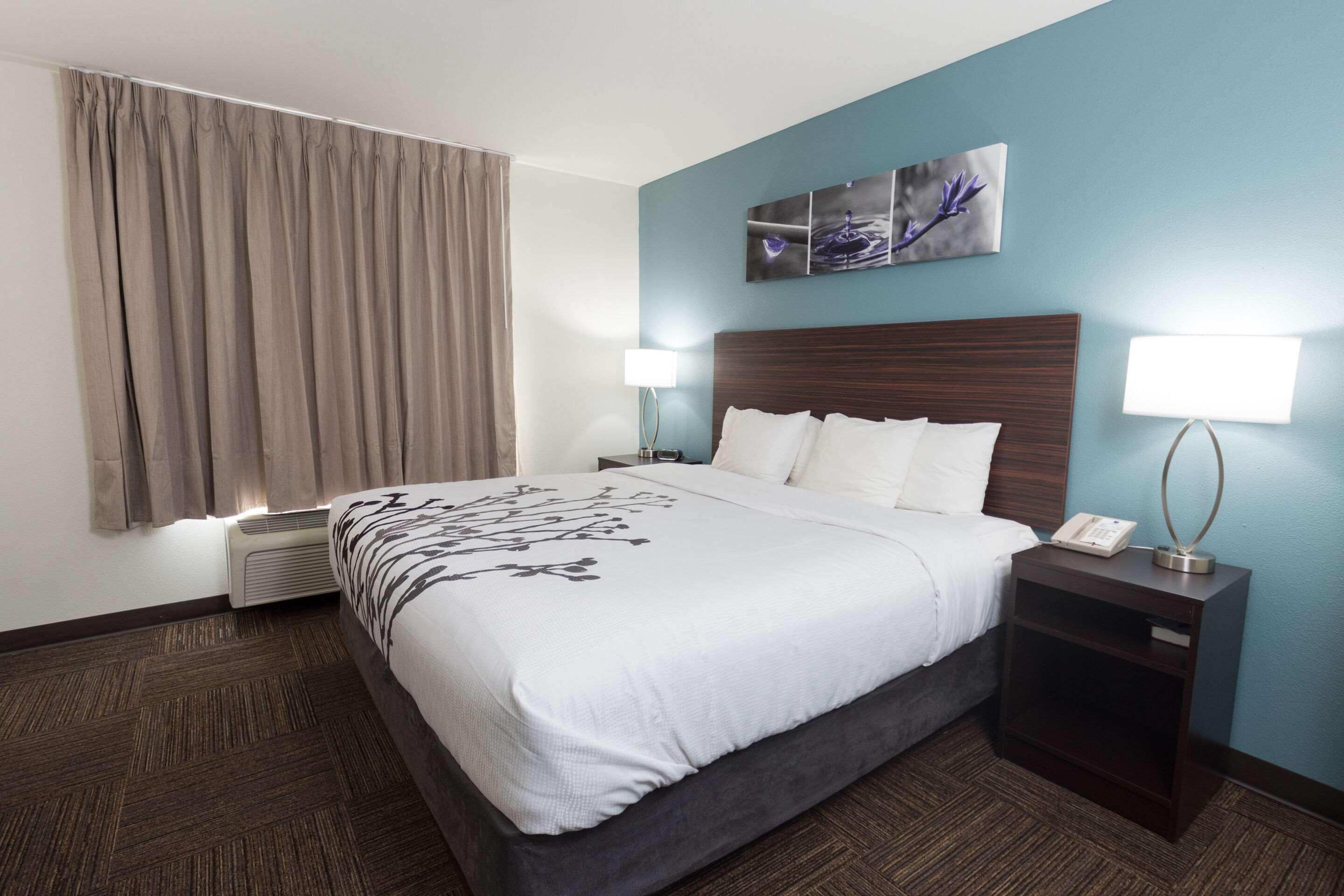 Quarto de hotel com uma cama grande, uma mesa de cabeceira com abajur, ar condicionado na parede e uma janela com cortinas.