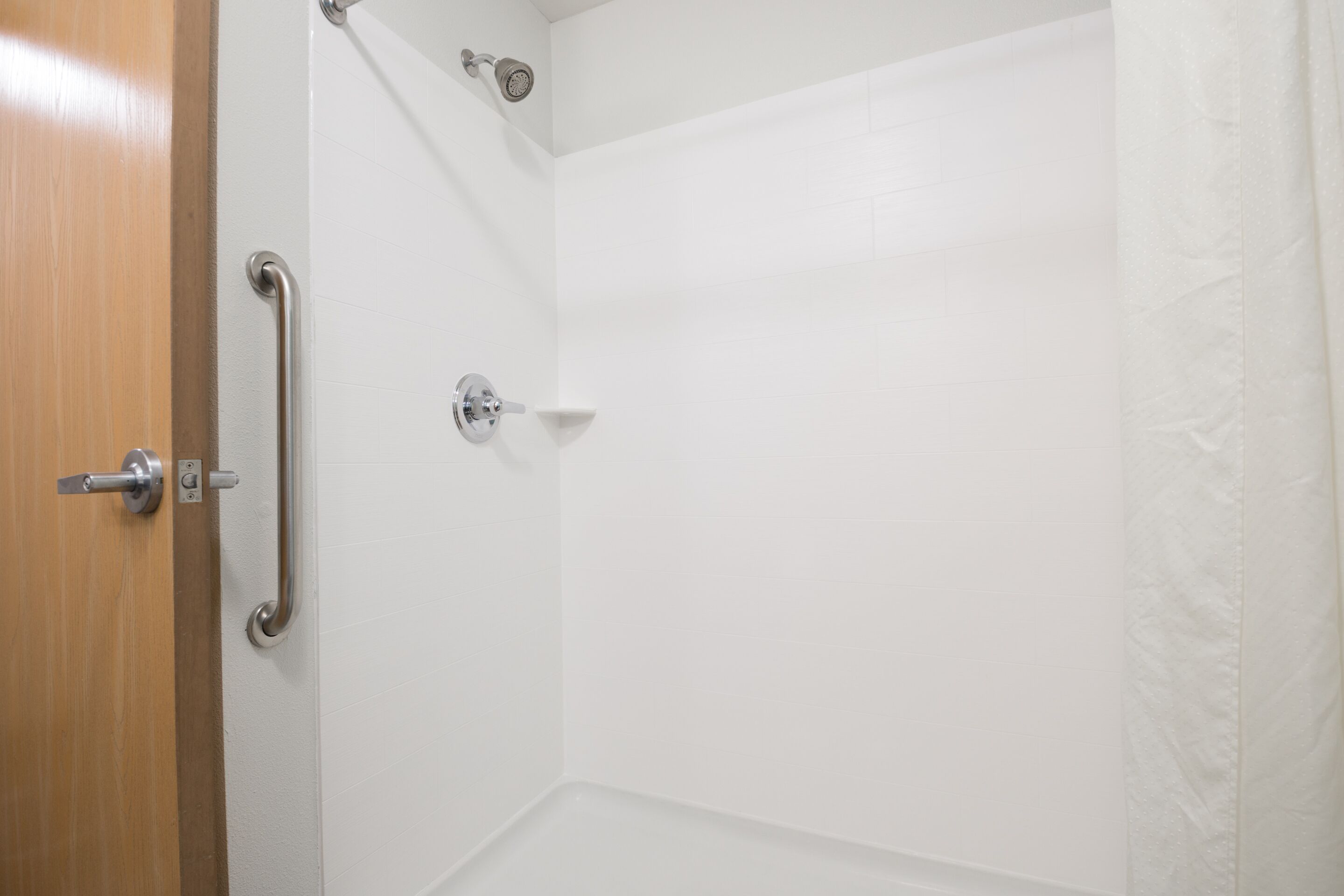A bathroom with a white tiled wall, a wooden door, a shower with a glass door, and a white shower curtain.