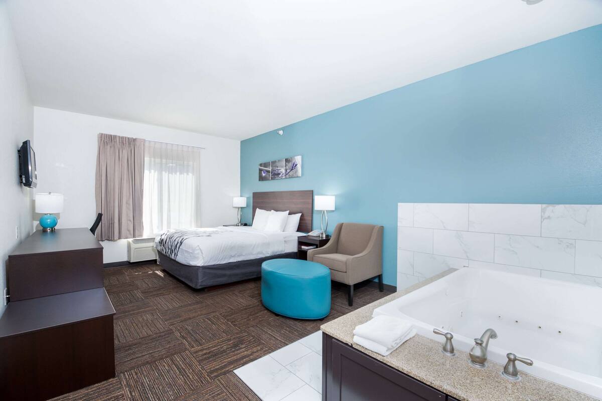 A hotel room with a bed, a desk, a chair, a blue ottoman, a large bathtub, and a TV.