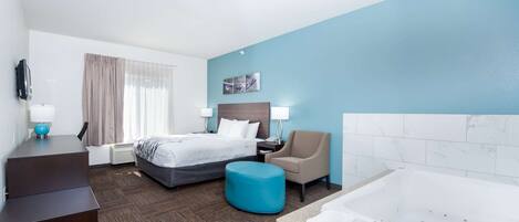 A hotel room with a bed, a desk, a chair, a blue ottoman, a large bathtub, and a TV.