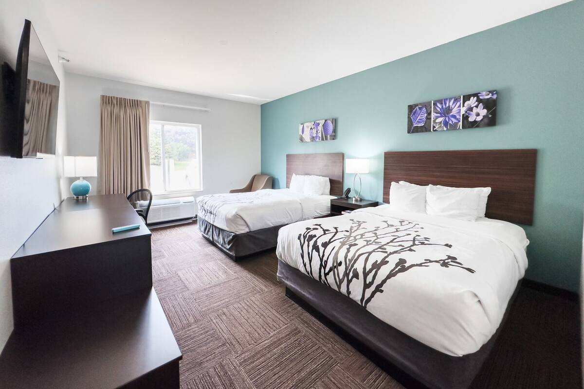 A hotel room with two beds, a desk, a chair, a TV, and artwork on the walls.