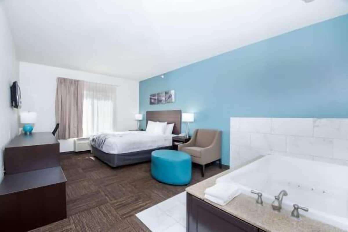 Quarto de hotel com cama, escrivaninha, cadeira, banheiro com banheira e uma grande janela com cortinas.