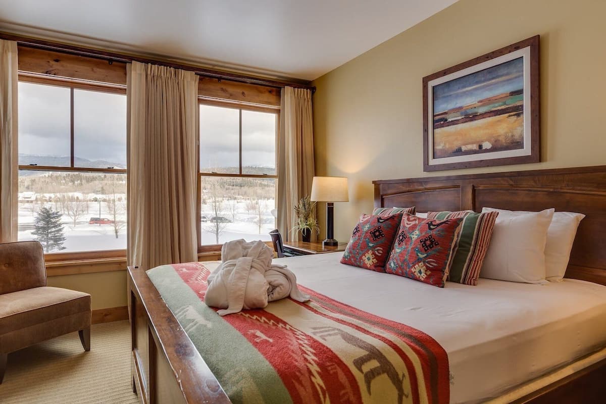 A hotel room with a large bed, a chair, a window with curtains, a painting on the wall, and a lamp on a bedside table.