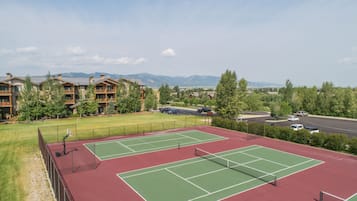 A tennis court with a net, surrounded by a fence, and a view of a lodge with multiple balconies and a parking area in the background.
