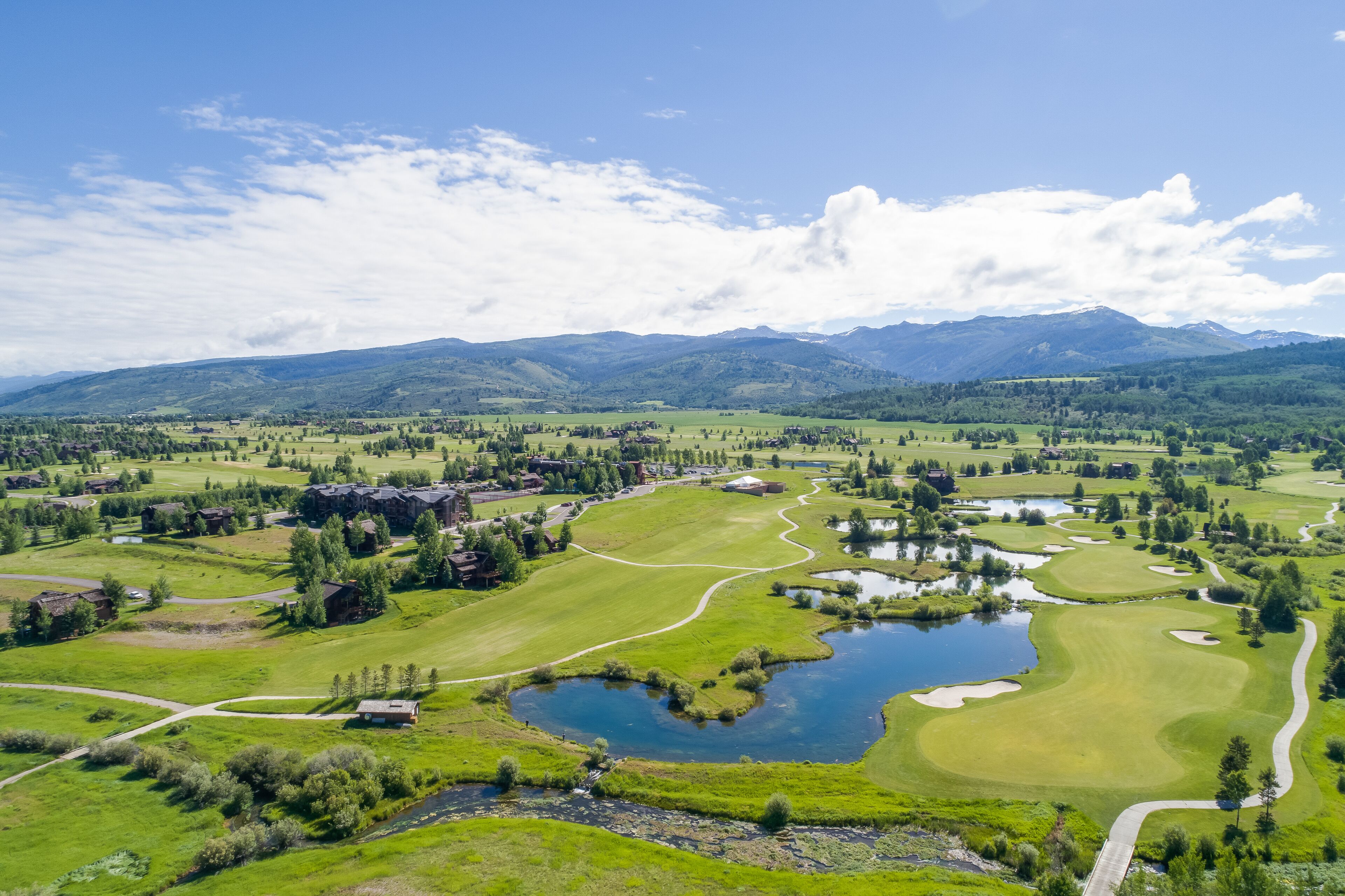A golf course with a pond, surrounded by green fields and mountains in the background.