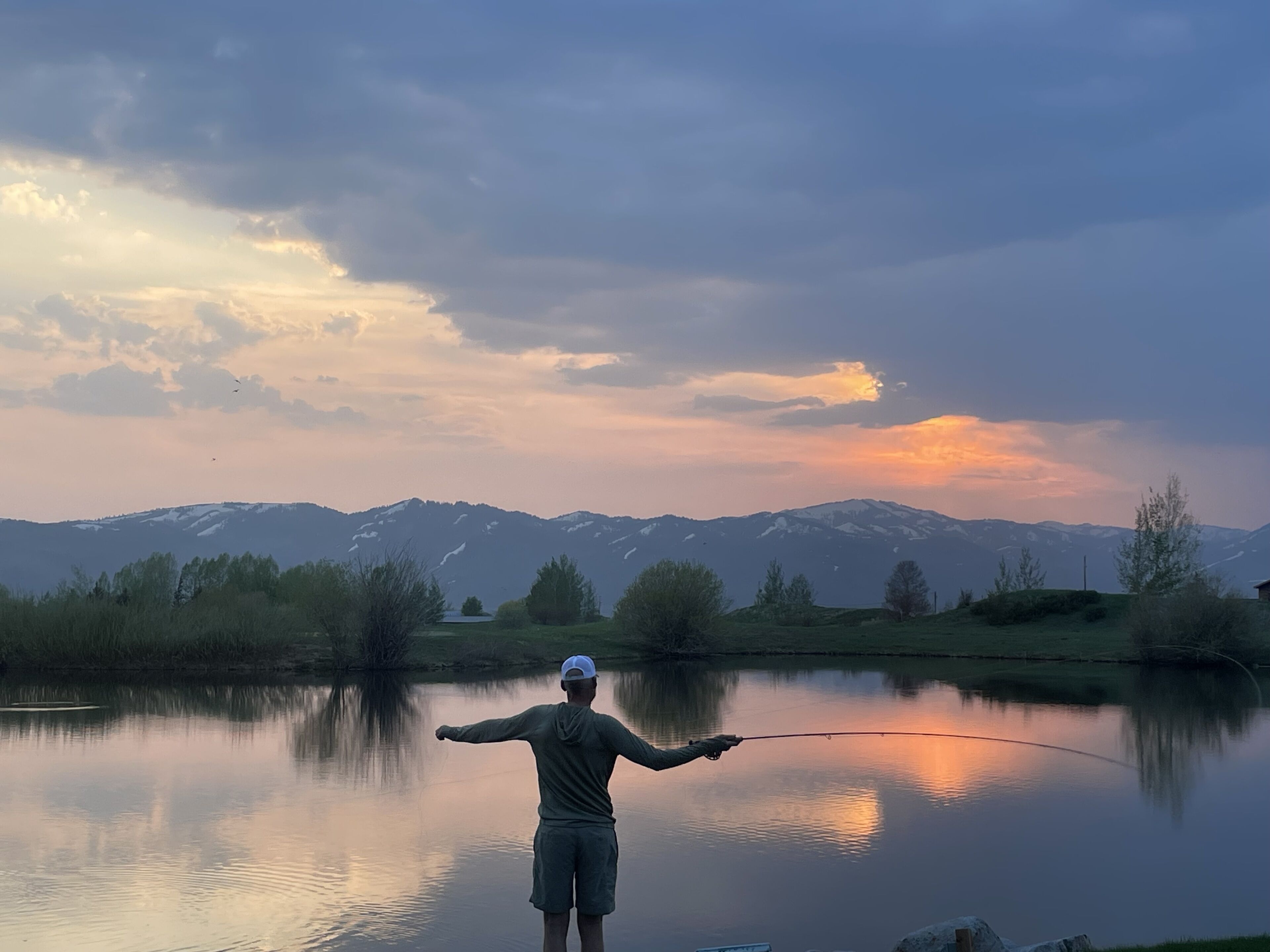 Alguém pesca num lago calmo, com montanhas ao fundo.