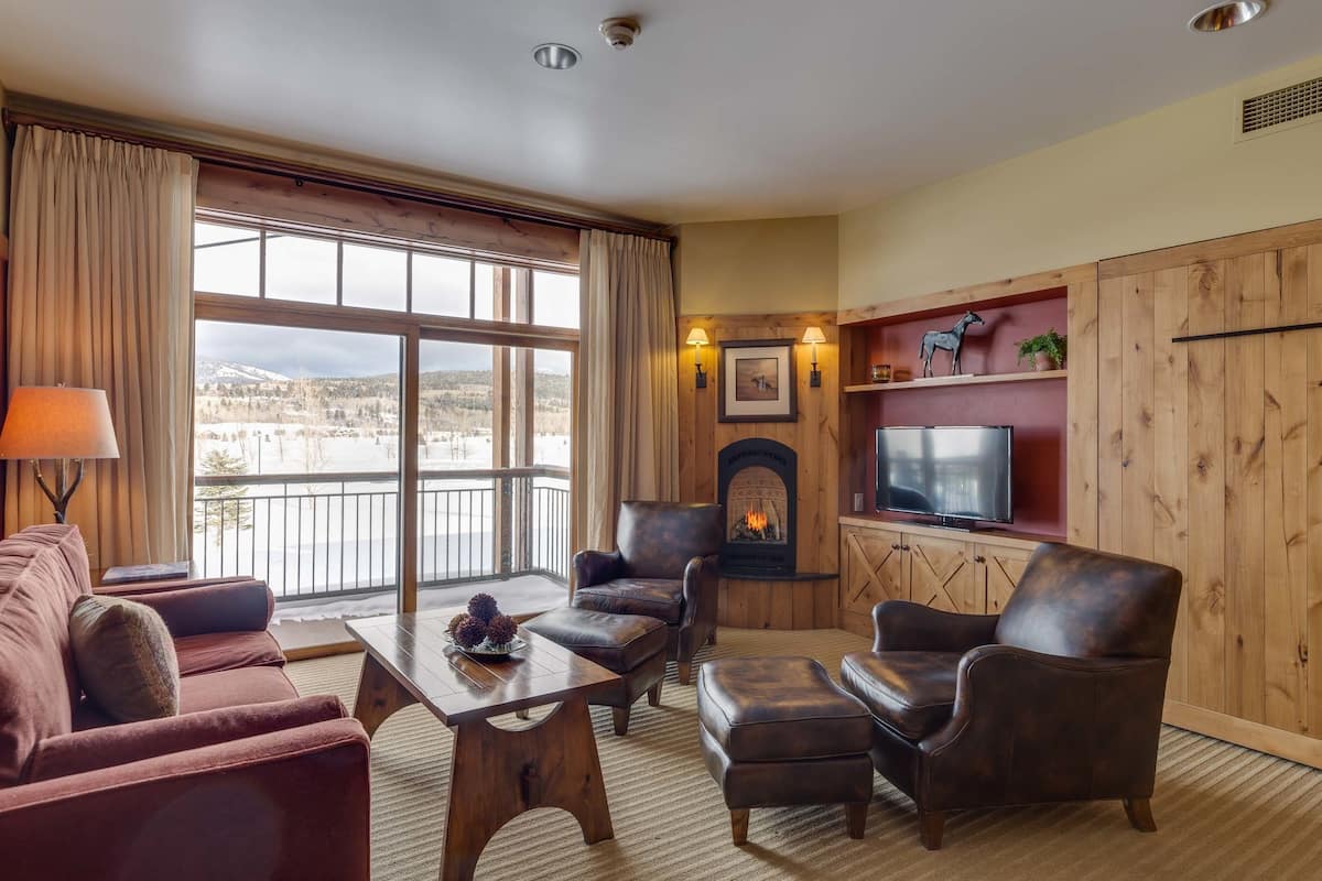 A cozy living room with leather furniture, a fireplace, a television, and a view of a snowy landscape through large windows.