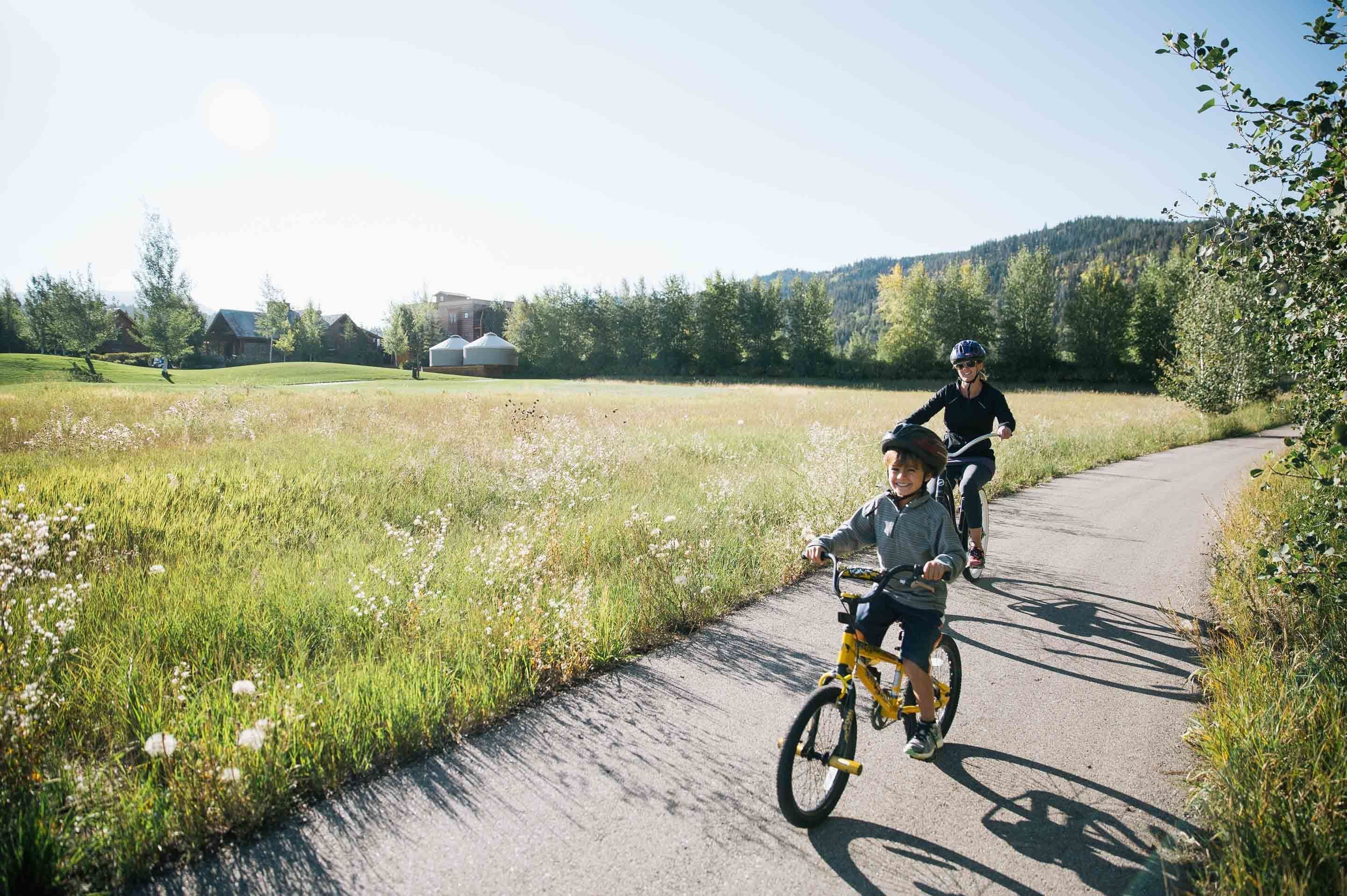 Two people riding bicycles on a paved path surrounded by greenery and a hilly landscape.
