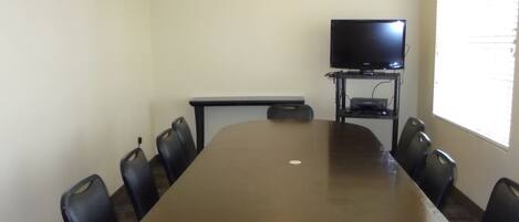 A conference room with a large table, eight chairs, a television on a stand, and a window with blinds.