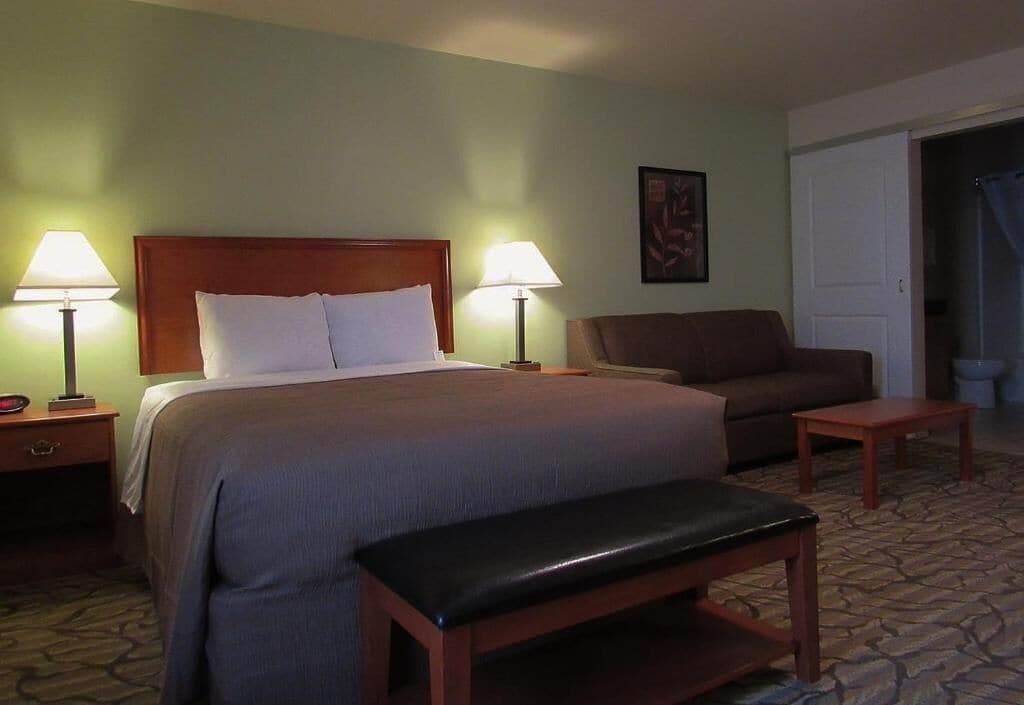 Quarto de hotel com cama, duas luminárias de cabeceira, um banquinho, uma cadeira e uma porta.
