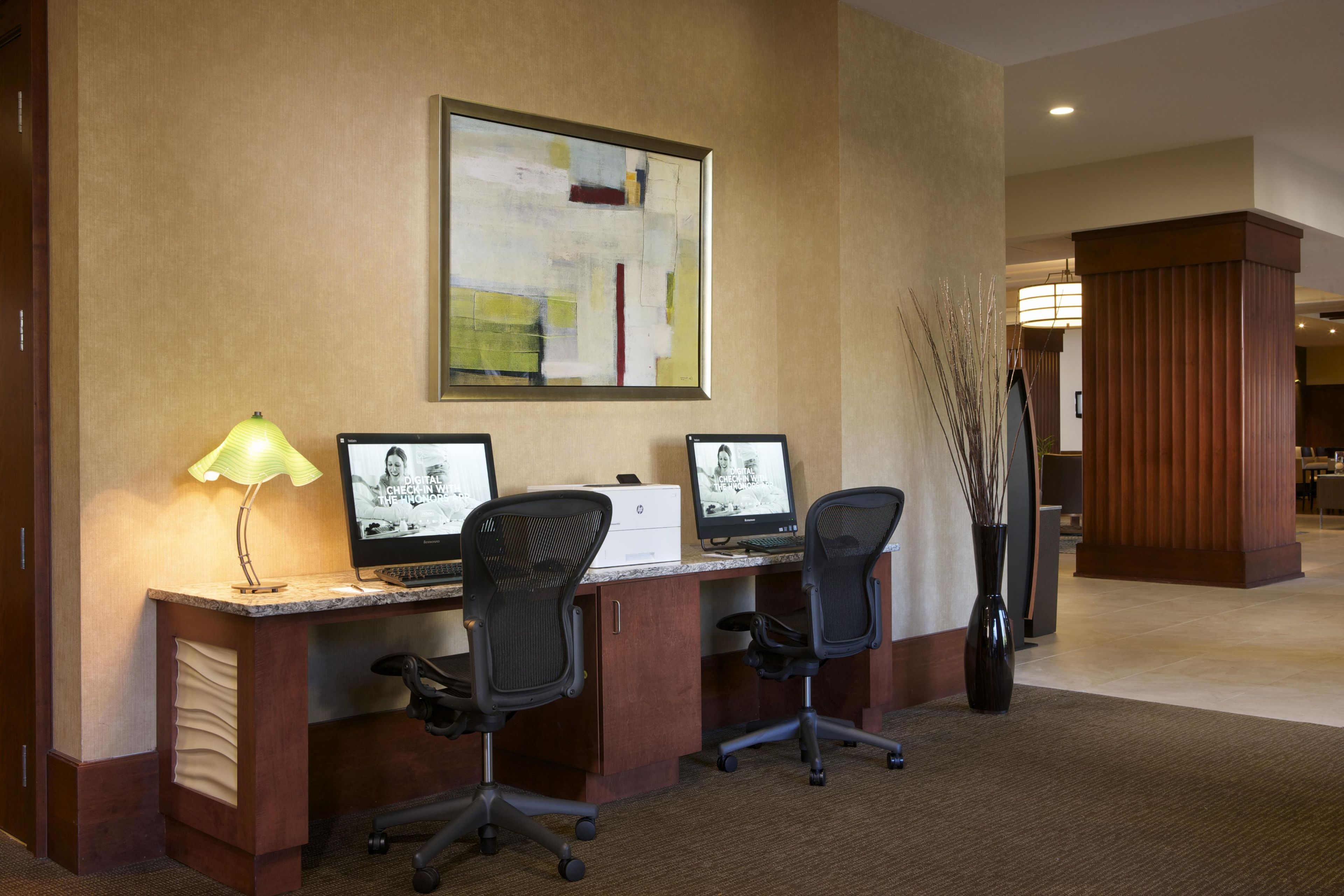 A hotel workspace with two desks, computers, chairs, a lamp, and a decorative wall piece.