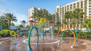 Un espace de jeux aquatiques avec des structures colorées, des palmiers et un bâtiment d’hôtel en arrière-plan.