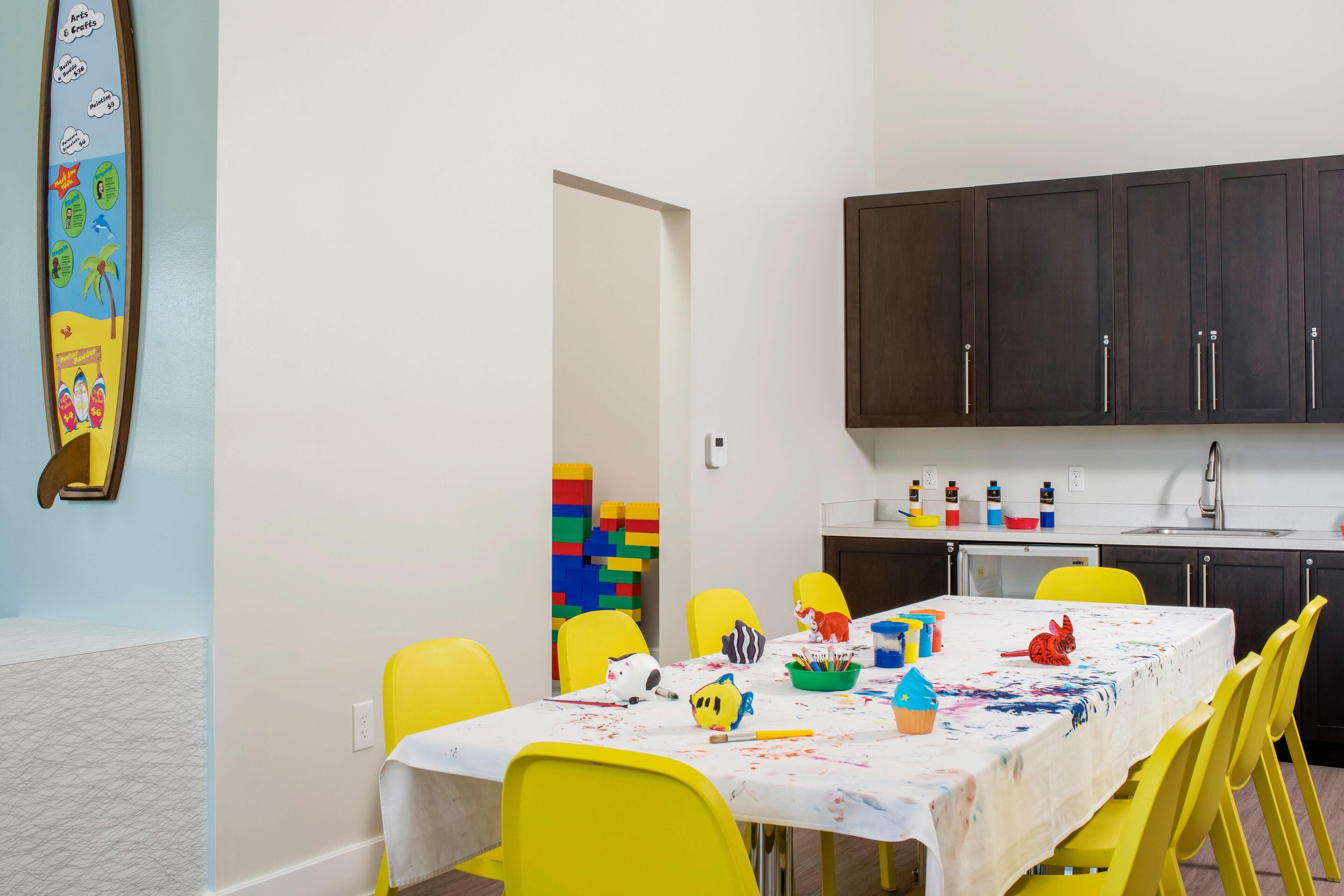 A dining area with a table covered in a white tablecloth, yellow chairs, and various children's toys and art supplies.