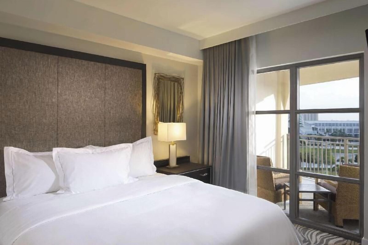 Een hotelkamer met een groot bed, een nachtkastje, een lamp, een raam met gordijnen en uitzicht op buiten.