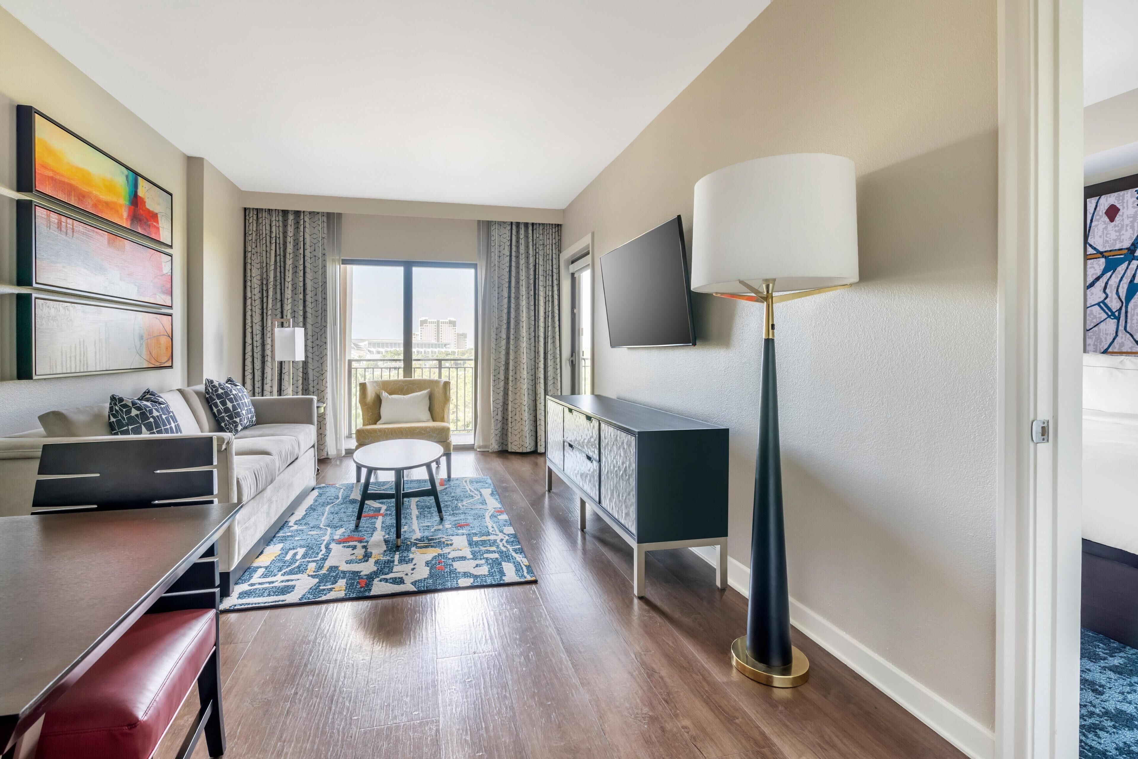A modern hotel room with a sofa, a dining table, a TV, and a floor lamp.