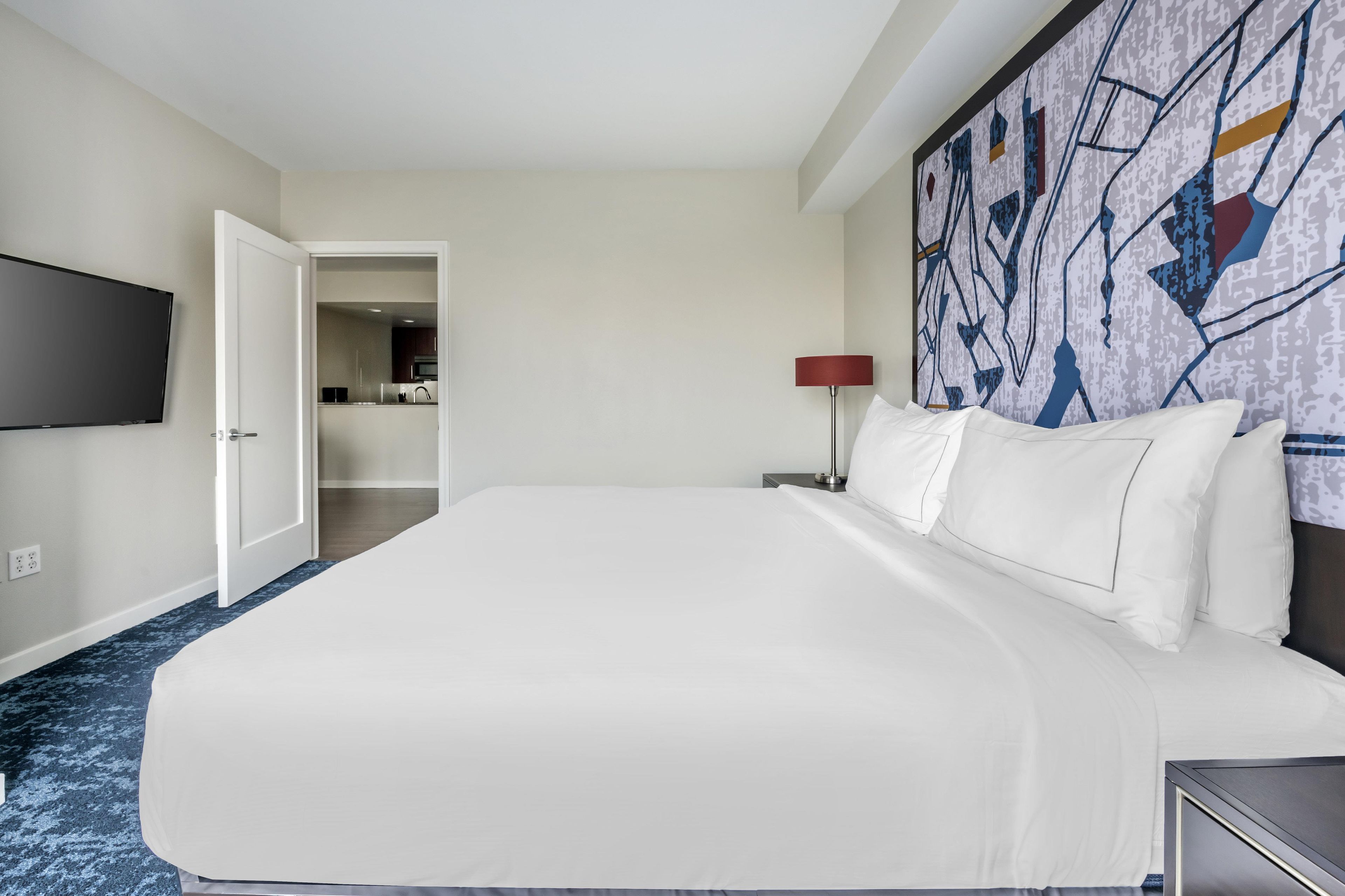 A hotel room with a large bed, a flat-screen TV mounted on the wall, and a modern abstract painting above the bed.