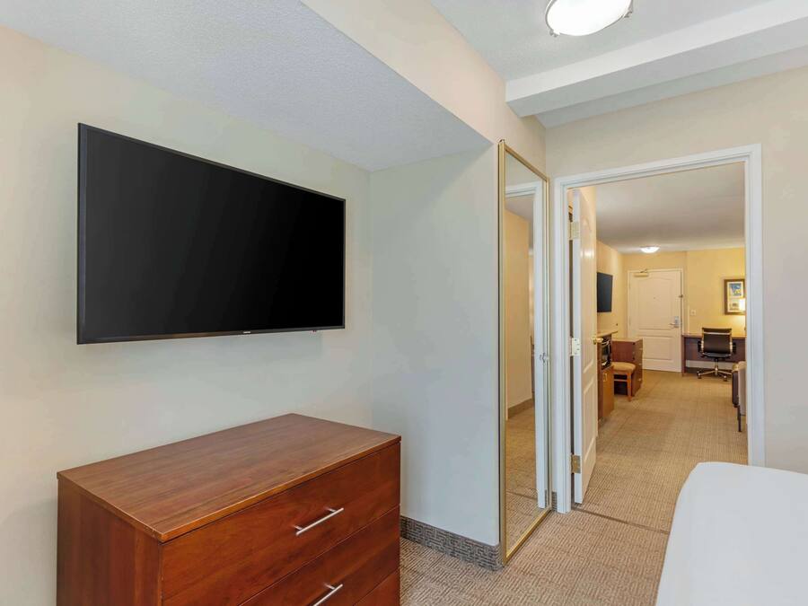 A hotel room with a flat-screen TV mounted on the wall, a wooden dresser, and a door leading to a bathroom and a small office area.