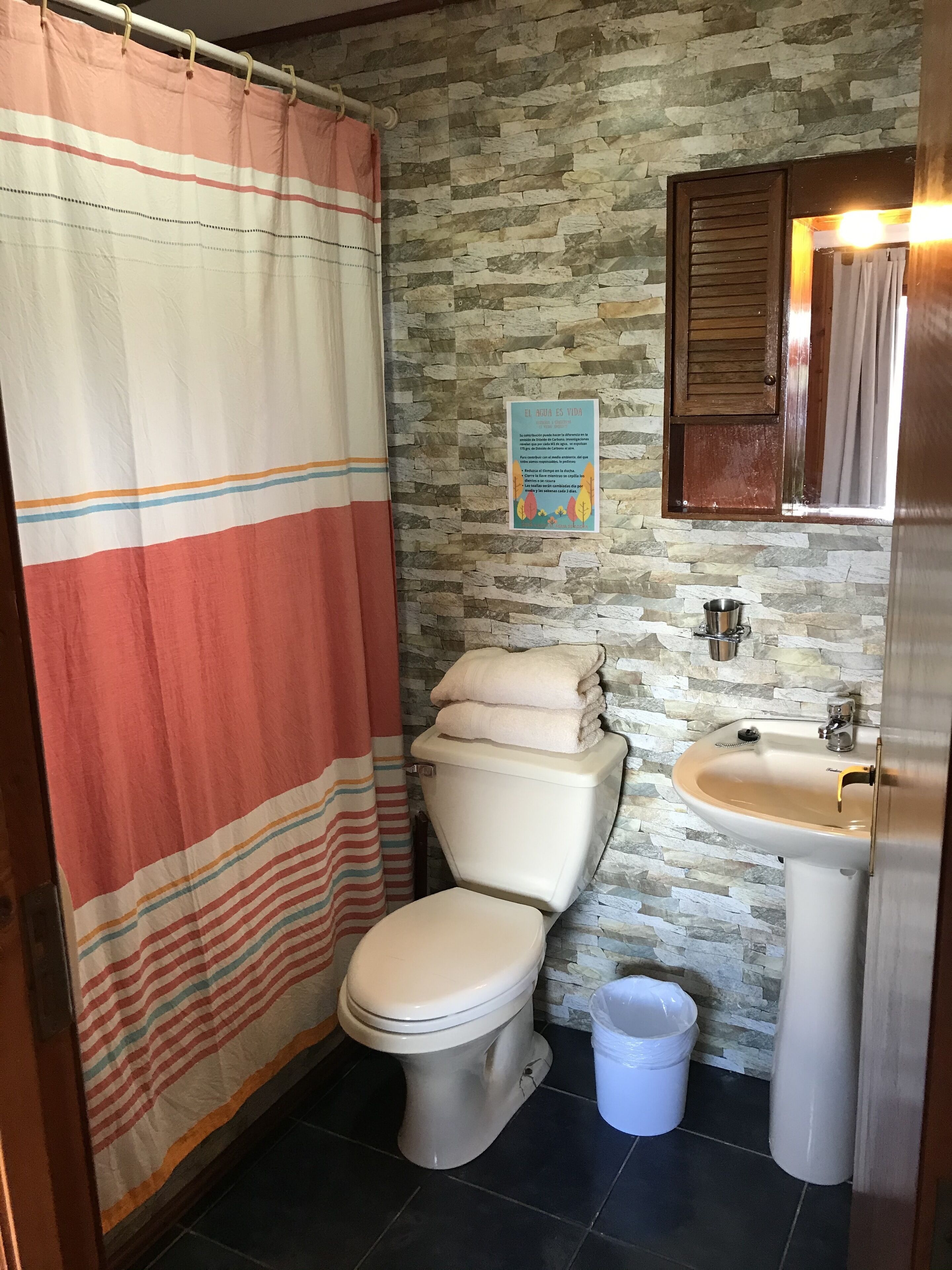 A bathroom with a striped shower curtain, a toilet, a sink, and a stone wall.