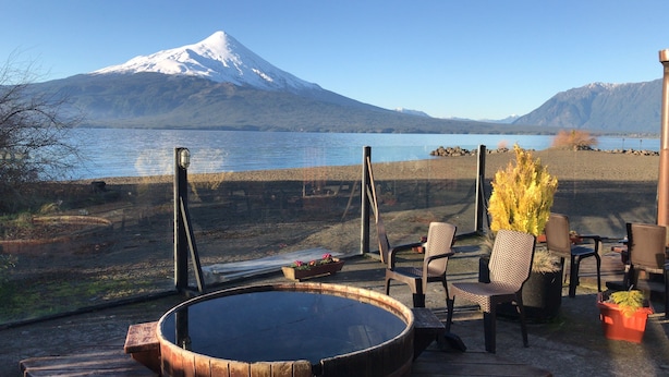 A wooden hot tub with chairs and a table, overlooking a lake with a mountain in the background. - Cabanas y Piscina Rucamalen (Puerto Varas)