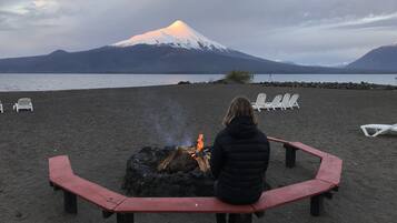 Una persona sentada en un banco en la playa, frente a un pozo de fuego, con vistas a una montaña y una extensión de agua.