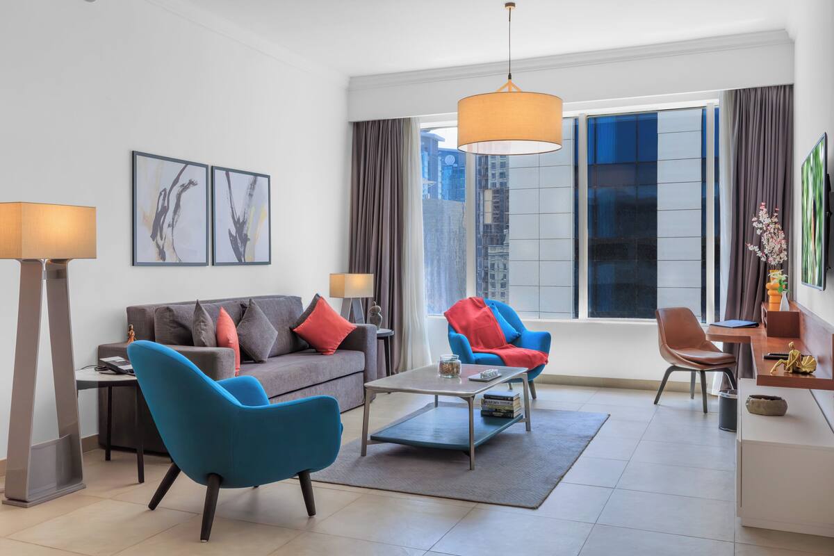 Ein modernes Wohnzimmer mit einer Couch, zwei Sesseln, einem Couchtisch, einer Lampe und Blick auf die Stadt durch große Fenster.
