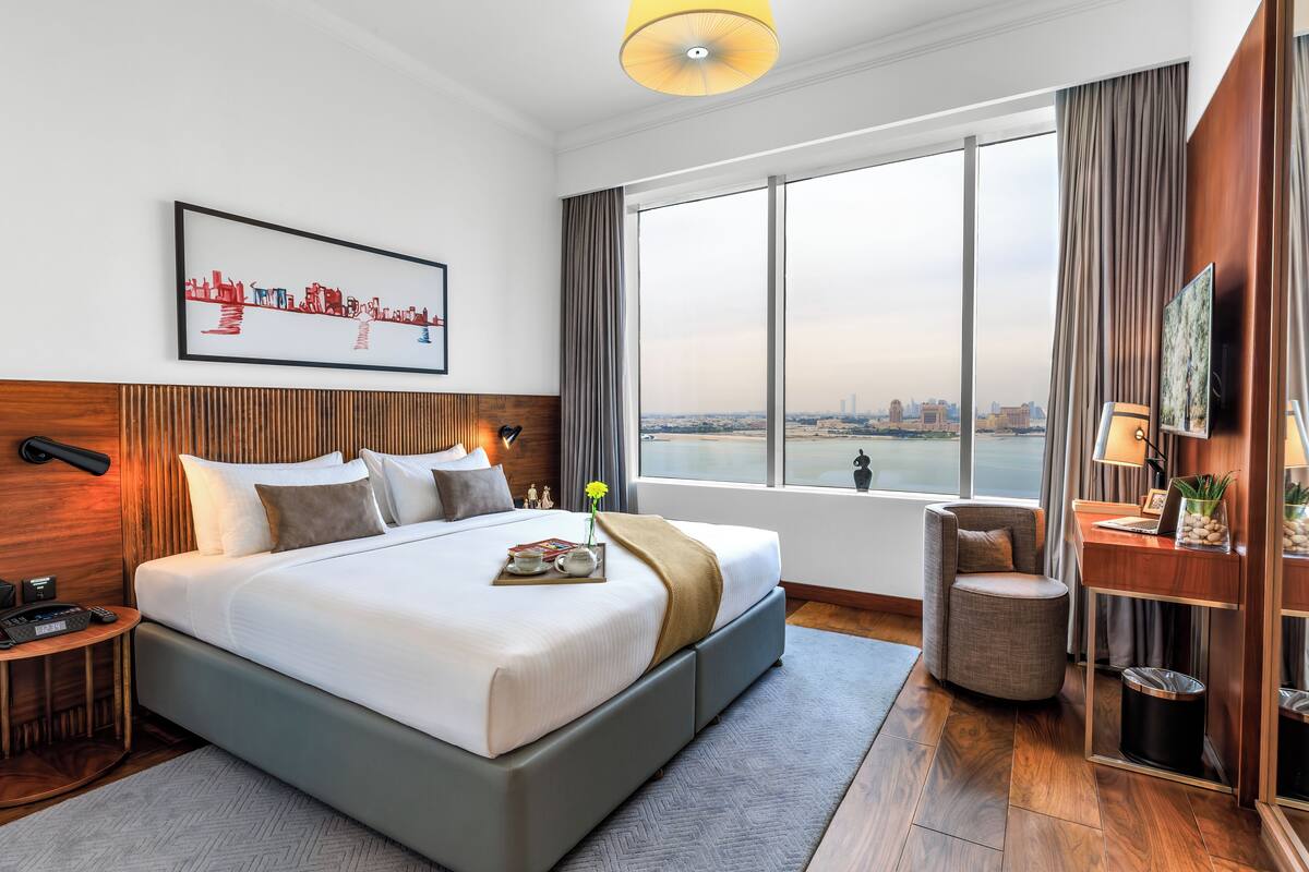 Ein Hotelzimmer mit einem großen Bett, einem Schreibtisch, einem Sessel, einem Fernseher und einem Fenster mit Blick auf die Stadt.