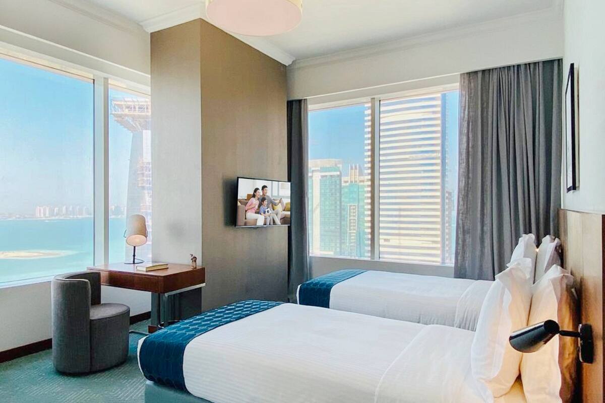 Ein Hotelzimmer mit zwei Betten, einem Schreibtisch, einem Sessel, einem Fernseher und Blick auf die Stadt.
