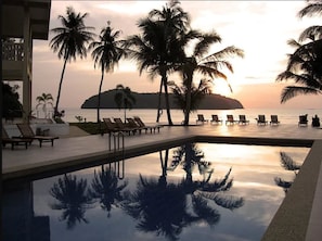 Una piscina con sillones de descanso y palmeras, reflejando el atardecer y una isla al fondo.