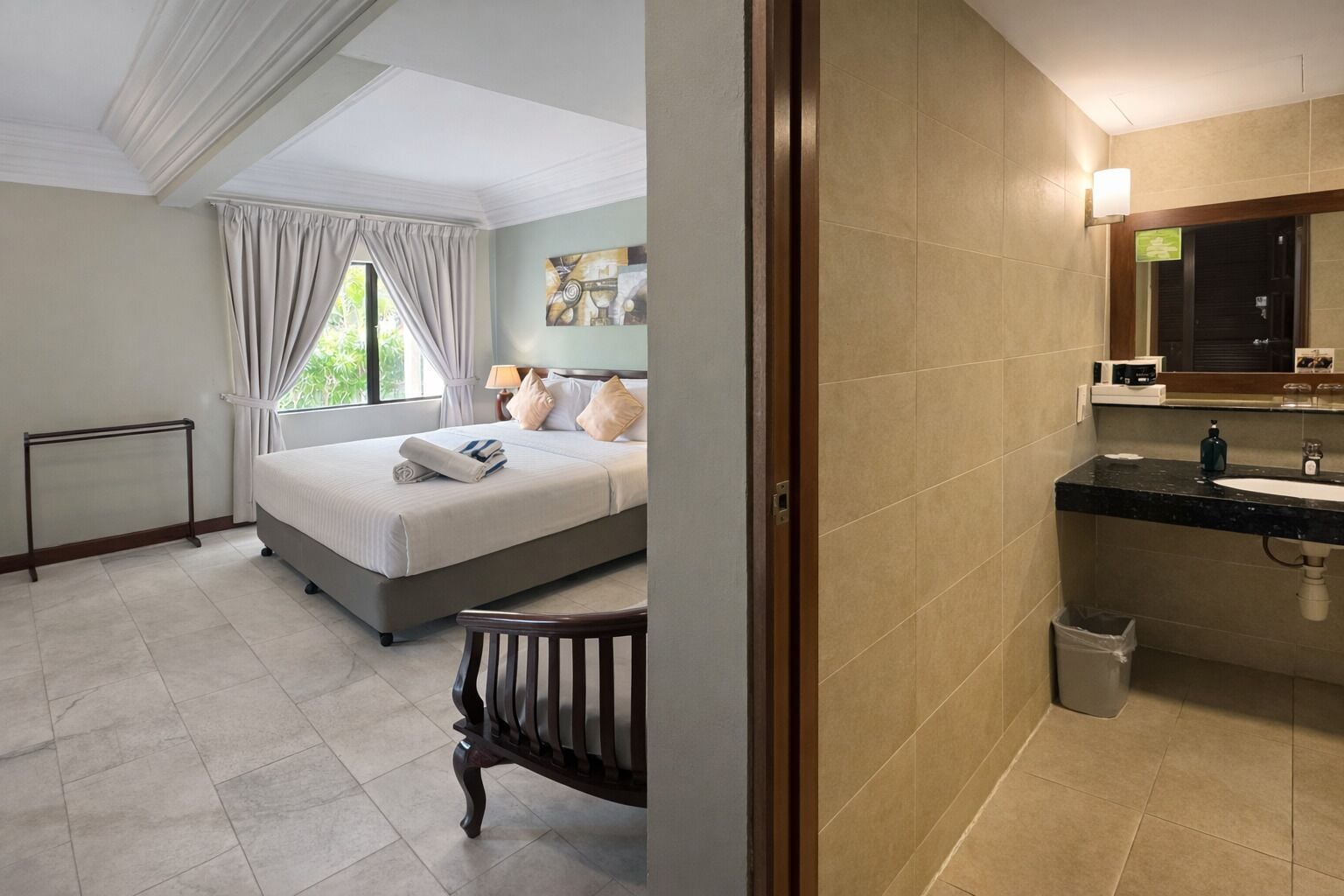 Ein Hotelzimmer mit Bett, Fenster mit Vorhängen, Badezimmer mit Waschbecken und Spiegel sowie ein Heizkörper.