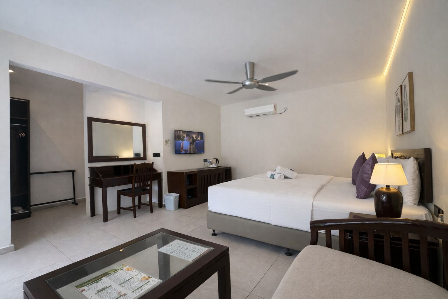 A hotel room with a bed, a desk, a chair, a television, a ceiling fan, and a nightstand with a lamp.