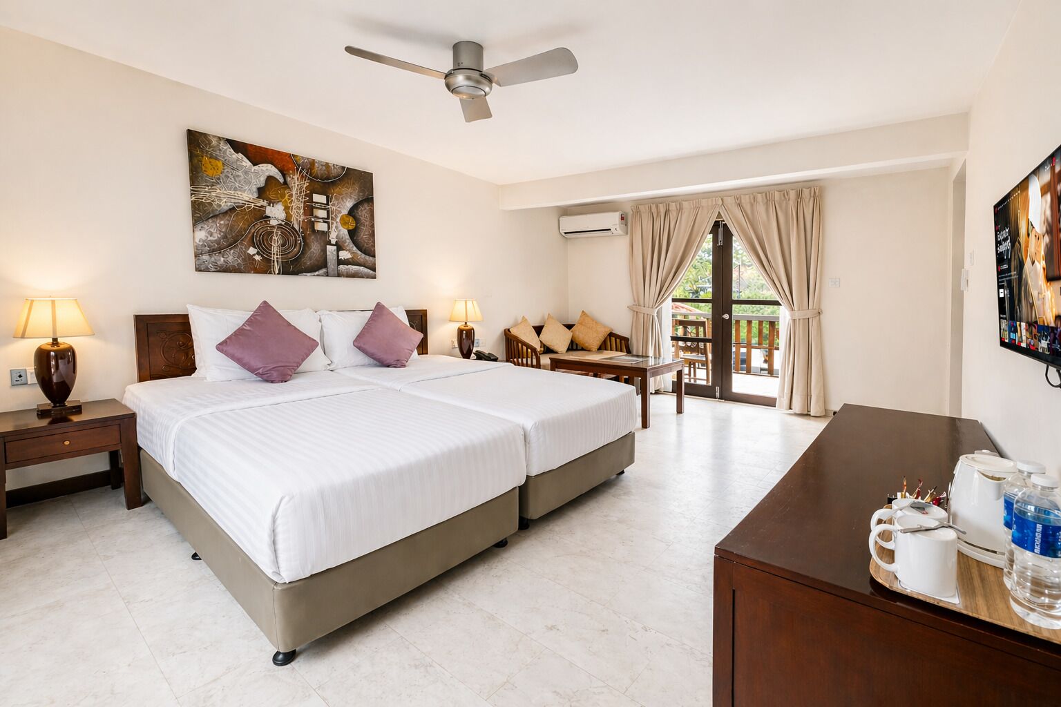 A hotel room with a bed, two bedside tables, a desk, a chair, a ceiling fan, an air conditioning unit, a painting, and a television.