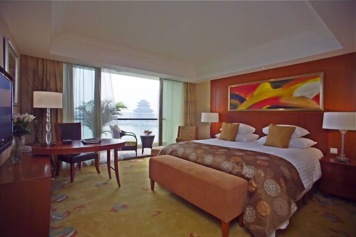 Habitación de hotel con una cama grande, un escritorio con silla, una computadora portátil, un televisor y vistas a un edificio tradicional.